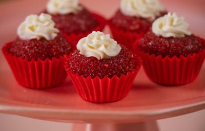 Fotografia de cinco unidades forminhas de cor vermelha, um junto da outra, e dentro de cada forminha tem um brigadeiro de red velvet, passado no açúcar cristal com uma decoração de cream cheese. Os brigadeiros estão sobre um apoio decorativo rosa alto. Fotografia de cinco unidades forminhas de cor vermelha, um junto da outra, e dentro de cada forminha tem um brigadeiro de red velvet, passado no açúcar cristal com uma decoração de cream cheese. Os brigadeiros estão sobre um apoio decorativo rosa alto.