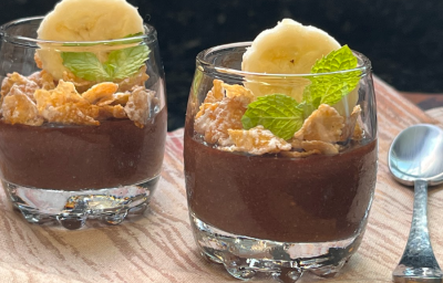 Imagem mostra 2 copinhos transparentes com um creme de chocolate escuro até a metade, seguido por cereais matinais, uma rodela de banana e folhas de hortelã. Os copos estão sobre um pano marrom e uma colher prateada está ao lado direito.
