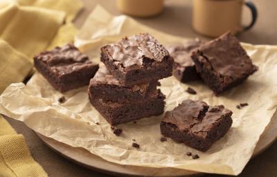 Fotografia em tons de marrom e bege em uma bancada de madeira, um pano bege, uma tábua de madeira redonda, um papel manteiga marrom com vários brownies de chocolate em cima. Ao fundo, xícaras beges. Fotografia em tons de marrom e bege em uma bancada de madeira, um pano bege, uma tábua de madeira redonda, um papel manteiga marrom com vários brownies de chocolate em cima. Ao fundo, xícaras beges.