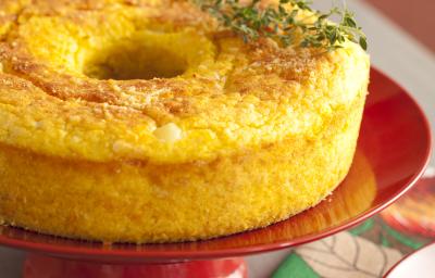 Fotografia em tons de cinza, vermelho e amarelo, de uma bancada cinza com um paninho estampado florido e um suporte de bolo vermelho, sobre ele um bolo de milho salgado e um ramo de orégano fresco. Fotografia em tons de cinza, vermelho e amarelo, de uma bancada cinza com um paninho estampado florido e um suporte de bolo vermelho, sobre ele um bolo de milho salgado e um ramo de orégano fresco.