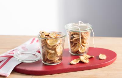 fotografia em tons de bege e vermelho tirada de uma bancada bege. Contém uma tábua vermelha com dois potes transparentes com bananas cortadas em rodelas finas fritas. Ao fundo um pano em tons de branco e vermelho. fotografia em tons de bege e vermelho tirada de uma bancada bege. Contém uma tábua vermelha com dois potes transparentes com bananas cortadas em rodelas finas fritas. Ao fundo um pano em tons de branco e vermelho.