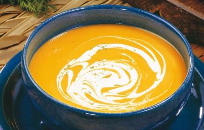 fotografia em tons de laranja, azul e marrom de uma bancada marrom vista de cima, contém um prato redondo azul com um recipiente redondo e azul com a sopa cremosa de cor laranja.