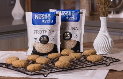 Receita de cookies proteicos servidos em uma grade sobre uma mesa de madeira com embalagens de Nestlé Aveia Proteína