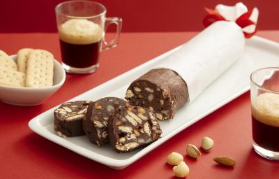 Foto da receita de Choco Salame. Observa-se uma cenografia vermelha com uma tábua branca e o chocosalame por cima, envolvido num papel anteiga. Dois copinhos de espresso decoram a foto. Foto da receita de Choco Salame. Observa-se uma cenografia vermelha com uma tábua branca e o chocosalame por cima, envolvido num papel anteiga. Dois copinhos de espresso decoram a foto.