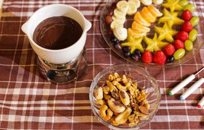 Fotografia em tons de marrom em uma bancada com uma toalha xadrez em marrom, com uma pequena panela branca com o fondue de chocolate dentro, ao fundo um recipiente com frutas picadas e um potinho de vidro com castanhas e amêndoas. Fotografia em tons de marrom em uma bancada com uma toalha xadrez em marrom, com uma pequena panela branca com o fondue de chocolate dentro, ao fundo um recipiente com frutas picadas e um potinho de vidro com castanhas e amêndoas.