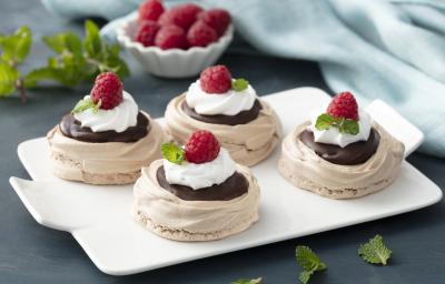 Fotografia em tons de vermelho em uma bancada de madeira azul escura, um pano azul claro, uma tábua branca ao centro com quatro Pavlovas em cima dela, decoradas com framboesa, chocolate e chantilly. Fotografia em tons de vermelho em uma bancada de madeira azul escura, um pano azul claro, uma tábua branca ao centro com quatro Pavlovas em cima dela, decoradas com framboesa, chocolate e chantilly.