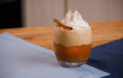 Copo baixo com bebida em camadas, espuma no topo, chantilly e canela em pau sobre tecido claro.