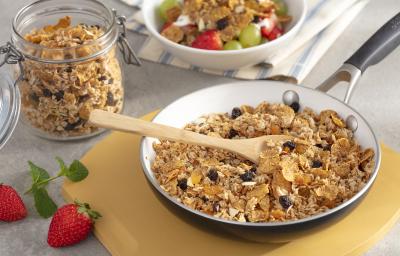 Fotografia em tons de amarelo em uma bancada de madeira com uma tábua de madeira, uma frigideira grande dentro com a granola com uvas-passas, cereal matinal dentro. Ao lado, um potinho com a granola e ao lado um outro potinho branco com frutas. Fotografia em tons de amarelo em uma bancada de madeira com uma tábua de madeira, uma frigideira grande dentro com a granola com uvas-passas, cereal matinal dentro. Ao lado, um potinho com a granola e ao lado um outro potinho branco com frutas.
