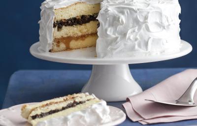 Fotografia em tons de branco e azul, ao fundo, sobre bancada azul, uma boleira de porcelana com bolo fatiado coberto com marshmallow e recheio de ameixa, prato de sobremesa com bolo de ameixa, dois pratos com talheres e um guardanapo com uma espátula.