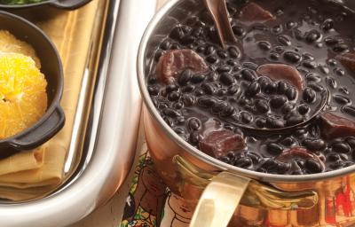 Fotografia em tons de bege em uma bancada de madeira clara com um suporte para panela colorido com rostos de mulheres, uma panela dourada em cima dele com a feijoada dentro.