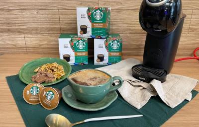 Fotografia em tons de verde e bege em uma bancada de madeira, um paninho bege, uma caneca verde com o petit gateau com café dentro. Ao fundo, máquina dolce gusto, e cápsulas de dolce gusto house blend. Fotografia em tons de verde e bege em uma bancada de madeira, um paninho bege, uma caneca verde com o petit gateau com café dentro. Ao fundo, máquina dolce gusto, e cápsulas de dolce gusto house blend.