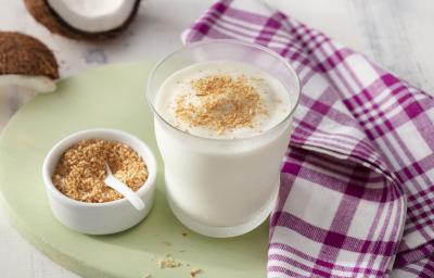 Fotografia em tons de branco, roxo e verde de uma bancada de madeira branca com uma tábua verde, sobre ela um paninho roxo e branco xadrez, um copo de vidro com a vitamina e um recipiente redondo branco com coco. Ao fundo um pedaço de coco fresco. Fotografia em tons de branco, roxo e verde de uma bancada de madeira branca com uma tábua verde, sobre ela um paninho roxo e branco xadrez, um copo de vidro com a vitamina e um recipiente redondo branco com coco. Ao fundo um pedaço de coco fresco.