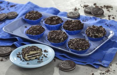 fotografia em tons de azul, preto e cinza de uma bancada cinza vista de cima, contém uma bandeja azul com forminhas azuis e brigadeiros de biscoitos e abaixo da bandeja um pano azul. Um pratinho na frente azul e redondo com uma fatia de brigadeiro.