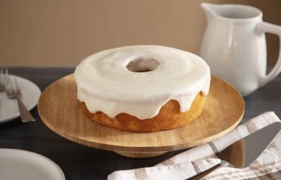 Foto em tons claros da receita de cobertura de porcelana de coco servida sobre um bolo em cima de uma base redonda de madeira. Ao lado há um prato de porcelana branco com talheres prateados e um pano bege à frente Foto em tons claros da receita de cobertura de porcelana de coco servida sobre um bolo em cima de uma base redonda de madeira. Ao lado há um prato de porcelana branco com talheres prateados e um pano bege à frente
