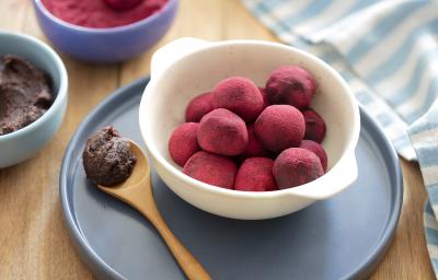 Fotografia em tons de roxo, em uma bancada de madeira marrom. Ao centro, uma tábua redonda azul e no meio um prato branco contendo as trufas. Ao lado, uma colher de madeira e ao fundo, há pó de beterraba em um prato. Fotografia em tons de roxo, em uma bancada de madeira marrom. Ao centro, uma tábua redonda azul e no meio um prato branco contendo as trufas. Ao lado, uma colher de madeira e ao fundo, há pó de beterraba em um prato.