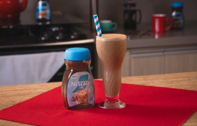Fotografia de um copo de vidro grande com shake de café, aveia e banana, ao lado de uma embalagem de nescafé gelado, sobre um pano vermelho.