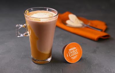 Fotografia em tons de laranja em uma bancada de madeira cinza escura, um paninho laranja e uma xícara de vidro alta com o café de mochaccino canela com doce de leite e achocolatado Galak.
