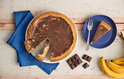 Fotografia em tons de azul em uma bancada de madeira, um pano azul, um prato grande raso com a cheesecake de banana com chocolate em cima dele. Ao lado, um prato pequeno azul raso com uma fatia da cheesecake, um cacho de banana e quadradinhos de chocolate Fotografia em tons de azul em uma bancada de madeira, um pano azul, um prato grande raso com a cheesecake de banana com chocolate em cima dele. Ao lado, um prato pequeno azul raso com uma fatia da cheesecake, um cacho de banana e quadradinhos de chocolate