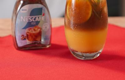 Fotografia de um copo de vidro com uma bebida de café gelado com tônica e laranja. A bebida está decorada com alecrim, e está ao lado de uma embalagem de nescafé gelado, sobre um pano vermelho.