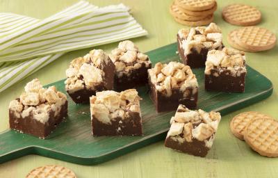 Fotografia em tons de verde de uma bancada verde com uma tábua verde, sobre ela pedaços de fudge. Ao fundo um paninho listrado verde e branco e biscoitos tostines. A frente biscoitos tostines. Fotografia em tons de verde de uma bancada verde com uma tábua verde, sobre ela pedaços de fudge. Ao fundo um paninho listrado verde e branco e biscoitos tostines. A frente biscoitos tostines.