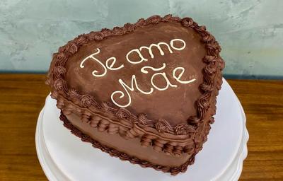 Fotografia em tons de castanho com um prato branco ao centro. Em cima do prato existe um bolo alto em formato de coração com cobertura de chocolate. Em cores brancas existe um escrito "Te amo Mãe" Fotografia em tons de castanho com um prato branco ao centro. Em cima do prato existe um bolo alto em formato de coração com cobertura de chocolate. Em cores brancas existe um escrito "Te amo Mãe"