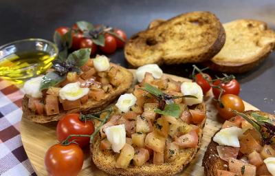 Foto da receita de brusqueta fácil em tons de bege servida em 3 porções dispersas em uma tábua de madeira oval sobre uma mesa quadriculada de cantina. Ao fundo, algumas fatias de pão italiano, tomates e em um recipiente com azeite para decorar
