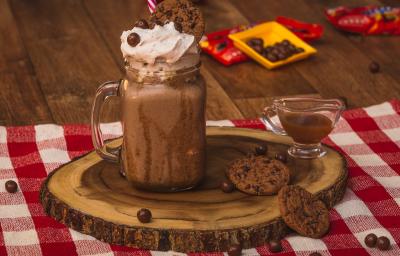 Foto de uma bancada de madeira. Sobre ela há uma toalha quadriculada vermelho e branco, uma tábua de madeira com um copo com o Milkshake de Nescau decorado com um canudo vermelho. Ao lado há um potinho com calda, 2 cookies e algunmas bolinhas de chocolate Foto de uma bancada de madeira. Sobre ela há uma toalha quadriculada vermelho e branco, uma tábua de madeira com um copo com o Milkshake de Nescau decorado com um canudo vermelho. Ao lado há um potinho com calda, 2 cookies e algunmas bolinhas de chocolate