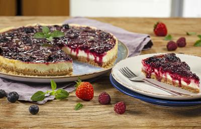 Fotografia em tons de vermelho em uma bancada de madeira com um prato raso grande com a cheesecake de iogurte com frutas vermelhas e um prato raso pequeno com uma fatia da cheesecake. Morangos e uvas espalhadas pela bancada. Fotografia em tons de vermelho em uma bancada de madeira com um prato raso grande com a cheesecake de iogurte com frutas vermelhas e um prato raso pequeno com uma fatia da cheesecake. Morangos e uvas espalhadas pela bancada.