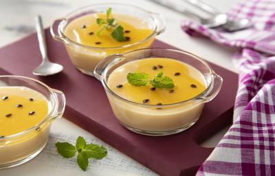 Fotografia em tons de amarelo, em uma bancada de madeira de cor branca. Ao centro, uma tábua de madeira roxa contendo 2 taças de mousse com uma colher ao lado. Ao fundo, há um pano quadriculado roxo. Fotografia em tons de amarelo, em uma bancada de madeira de cor branca. Ao centro, uma tábua de madeira roxa contendo 2 taças de mousse com uma colher ao lado. Ao fundo, há um pano quadriculado roxo.