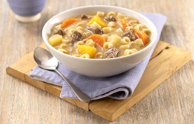 Foto em tons de amarelo suave com a sopa de macarrão, carne e legumes em um bowl branco. Há um paninho azul embaixo do bowl e em cima de uma tábua de madeira quadrada com uma colher prateada na frente. Todos esses elementos em cima de uma mesa de madeira.