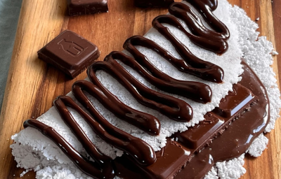 A imagem mostra uma tapioca branca dobrada ao meio, recheada e coberta com um creme de chocolate e pedaços de chocolate. A tapioca está apoiada em uma tábua de madeira com pedaços de chocolate espalhados.