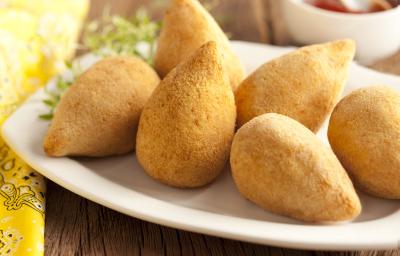 Fotografia em tons de amarelo em uma bancada de madeira, um pano amarelo, uma tábua de madeira, um prato branco oval com várias coxinhas de frango em cima dele. Ao fundo, um potinho com ketchup. Fotografia em tons de amarelo em uma bancada de madeira, um pano amarelo, uma tábua de madeira, um prato branco oval com várias coxinhas de frango em cima dele. Ao fundo, um potinho com ketchup.