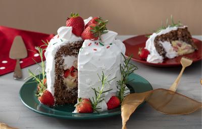 Fotografia em tons de branco em uma bancada de madeira de cor cinza. Ao centro, um prato verde contendo o panettone. Ao fundo, há um pires vermelho com uma fatia do panettone e uma toalha vermelha com uma espátula em cima. Fotografia em tons de branco em uma bancada de madeira de cor cinza. Ao centro, um prato verde contendo o panettone. Ao fundo, há um pires vermelho com uma fatia do panettone e uma toalha vermelha com uma espátula em cima.