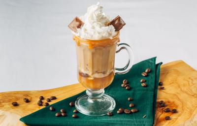 Fotografia em tons de marrom em uma bancada de madeira de cor branca com uma tábua de madeira. Em cima, há um pano verde contendo uma caneca com a bebida. Ao redor, há vários grãos de café espalhados.