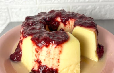 Fotografia em tons claros com um pudim de NINHO coberto com geleia de frutas vermelhas ao centro. O pudim possui cor clara e a geleia de frutas vermelhas escorre delicadamente sobre o pudim.