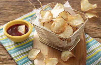 Fotografia em tons de amarelo e verde em uma bancada de madeira, com um pano listrado em amarelo e verde e uma tábua de madeira com uma cestinha de tela com várias batatas chips. Ao lado, um potinho amarelo com ketchup. Fotografia em tons de amarelo e verde em uma bancada de madeira, com um pano listrado em amarelo e verde e uma tábua de madeira com uma cestinha de tela com várias batatas chips. Ao lado, um potinho amarelo com ketchup.