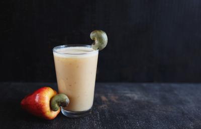 Fotografia de um copo de vidro com refresco de Neston com suco de caju, ao lado de um caju in natura sobre uma mesa escura e um fundo preto.