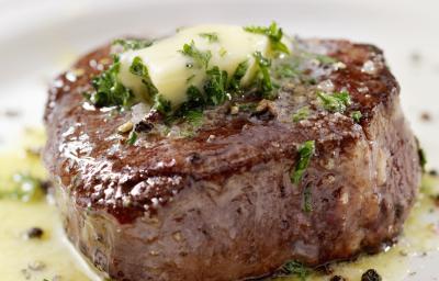 Fotografia de um bife mais alto feito, em um prato branco, com um azeite de ervas e pimenta sobre a carne e escorrendo no prato. Em cima da carne, manteiga e salsinha.