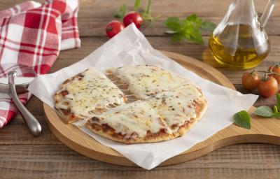 Fotografia em tons de amarelo e bege de uma bancada de madeira, ao centro uma tabua de madeira com um pedaço de papel manteiga e um disco de pizza com molho e queijo derretendo. Ao fundo um pano xadrez vermelho e branco e um vidro com azeite. Fotografia em tons de amarelo e bege de uma bancada de madeira, ao centro uma tabua de madeira com um pedaço de papel manteiga e um disco de pizza com molho e queijo derretendo. Ao fundo um pano xadrez vermelho e branco e um vidro com azeite.
