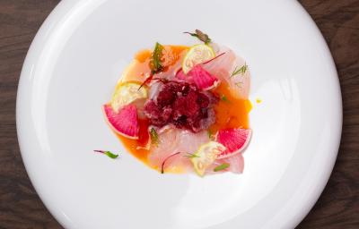 Fotografia em tons de branco com um prato branco ao centro. Em cima do prato existe uma receita de crudo de peixe de cor laranja e rosa, acompanhada de pedaços de limão Fotografia em tons de branco com um prato branco ao centro. Em cima do prato existe uma receita de crudo de peixe de cor laranja e rosa, acompanhada de pedaços de limão