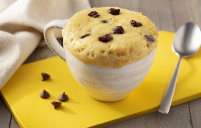 Fotografia em tons de marrom. branco e amarelo de uma bancada de madeira com uma tábua amarela, sobre ela uma xícara branca com bolo de caneca e uma colher. Ao fundo um paninho branco.
