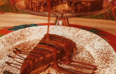Fotografia de uma fatia de torta de ninho e chocolate em um prato branco raso salpicado de cacau em pó. Por cima da fatia tem calda.