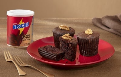 Fotografia de três muffins de nescau sobre um prato vermelho, raso, ao lado de dois garfos dourados e uma lata de NESCAU Extra Cacau. Um dos muffins está cortado na metade para mostrar a maciez da massa. Fotografia de três muffins de nescau sobre um prato vermelho, raso, ao lado de dois garfos dourados e uma lata de NESCAU Extra Cacau. Um dos muffins está cortado na metade para mostrar a maciez da massa.