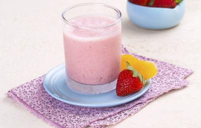 Fotografia em tons de branco e rosa de uma bancada cor branca vista de cima, ao centro um pano cor rosa e em cima um pratinha cor azul que contém um copo transparente com bebida de morango e ao lado um morango e uma fatia de laranja para decorar.