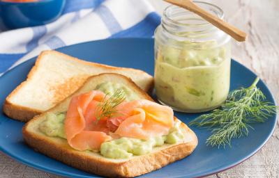 fotografia em tons de cinza, azul e verde tirada de uma bancada cinza, com um prato azul e redondo que contém 2 pães de forma e um deles contém salmão e guacamole. Ao lado esquerdo do prato contém um potinho com a guacamole dentro.