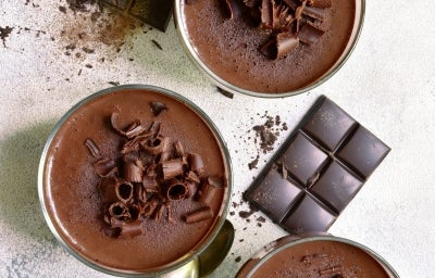 Fotografia vista de cima, em tons claros, de três recipientes de vidro sobre uma mesa clara. Dentro dos potinhos tem a sobremesa, finalizada com ganache e raspas de chocolate, e ao redor tem raspas e algumas barras pequenas de chocolate.