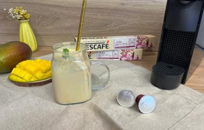 Fotografia em tons de amarelo com uma caneca ao centro. Dentro da caneca existe um líquido de cor bege com um ramo de hortelã de decoração. Ao fundo existe uma maquina de café, uma manga aberta e 2 caixas de cápsulas de café NESCAFÉ