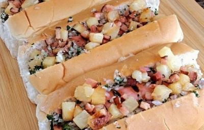 Foto em tons marrom claro da receita de cachorro quente havaiano servida em três porções uma ao lado da outra sobre uma tábua de madeira em cima de uma mesa de mármore Foto em tons marrom claro da receita de cachorro quente havaiano servida em três porções uma ao lado da outra sobre uma tábua de madeira em cima de uma mesa de mármore