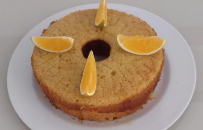Fotografia de um bolo de laranja feito na forma de fundo central, sobre um prato de vidro branco. Sobre o bolo tem quatro pedaços de laranja cortados. Fotografia de um bolo de laranja feito na forma de fundo central, sobre um prato de vidro branco. Sobre o bolo tem quatro pedaços de laranja cortados.