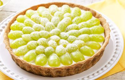 Fotografia em tons de verde e branco, prato branco ao centro contendo uma torta de uva sobre guardanapo amarelo, ao lado potinho com uvas, tudo sobre bancada de madeira com tons em branco